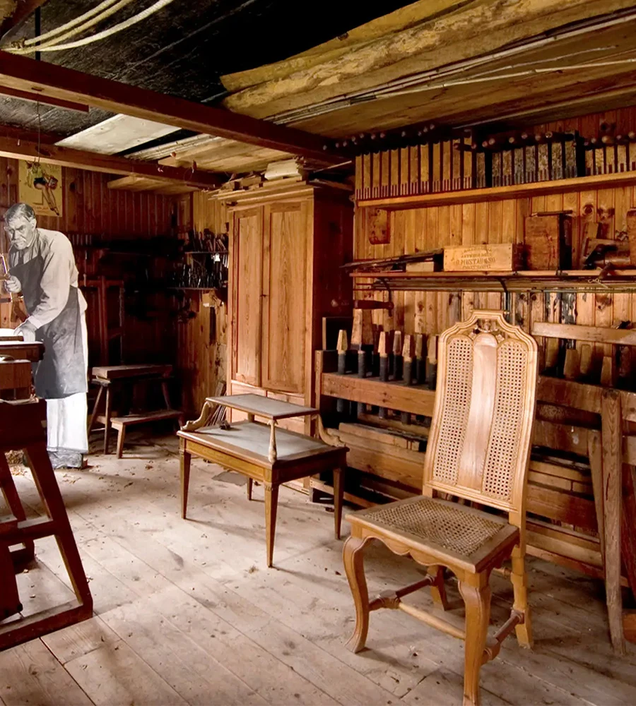 En verkstad med trämöbler och verktyg, i hörnet står en pappfigur av ett fotografi av möbelsnickare Forsman i naturlig storlek och sågar.