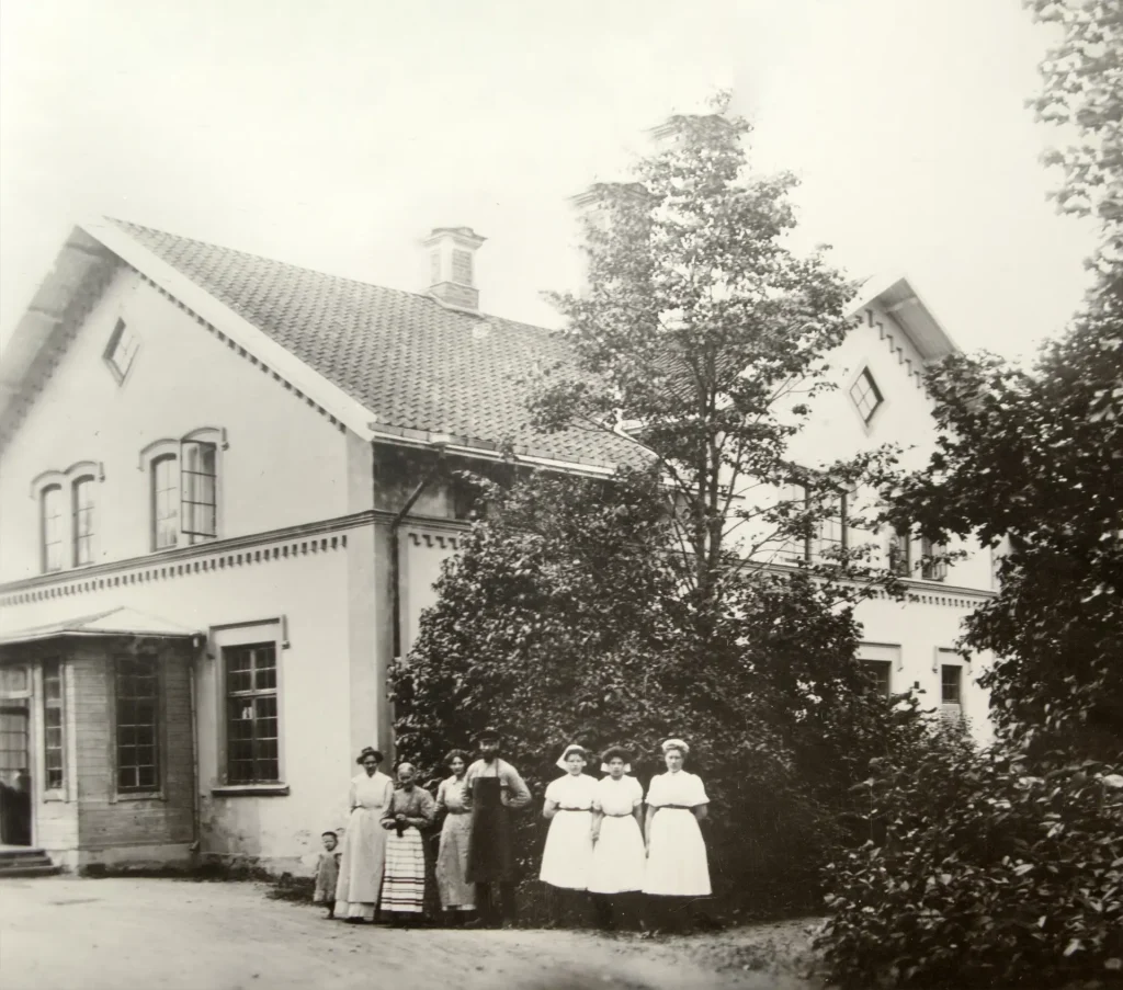 Svartvitt fotografi av ett putsat hus, framför står en grupp mejerskor i vita kläder, en man med hatt och skägg och läderförkläde.