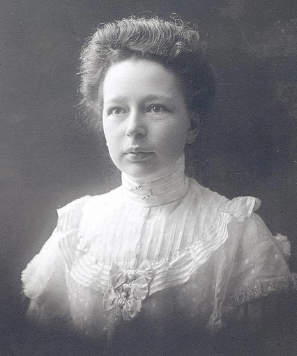 Porträtt på kvinna med uppsatt hår, hon bär en ljus kråsblus med hög hals och tre rosetter.
