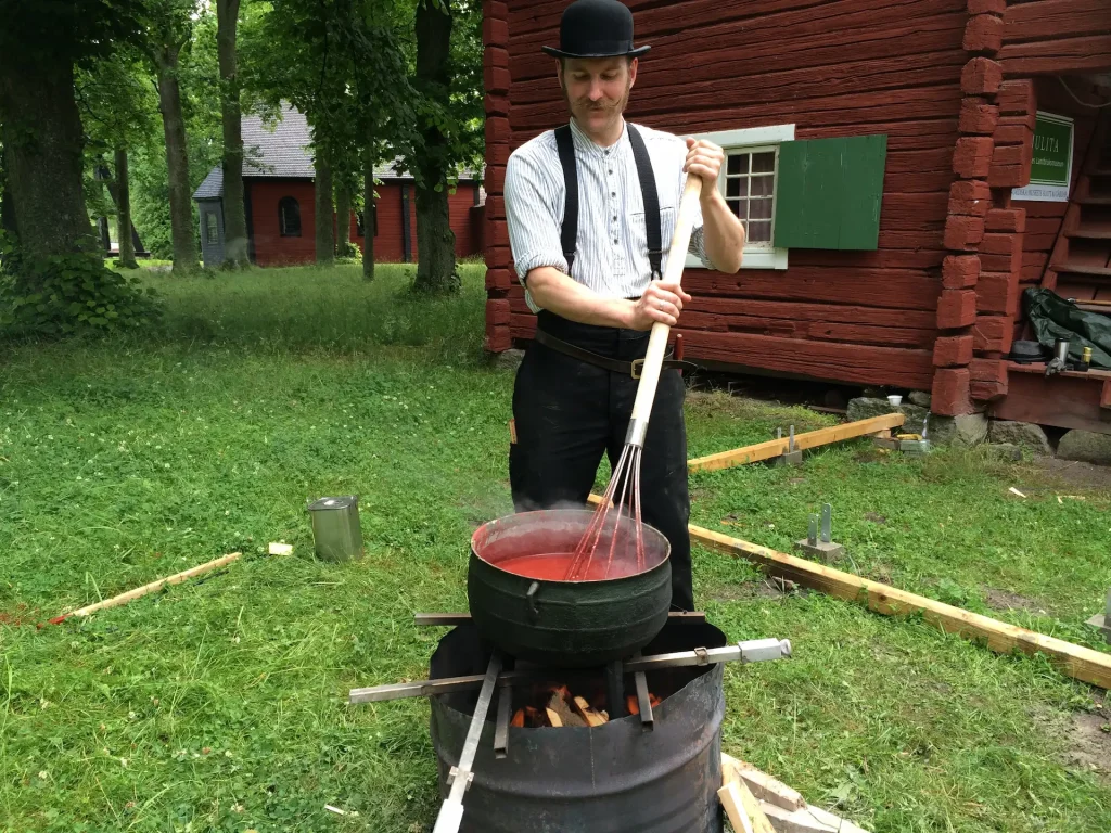 En man kokar färg utomhus klädd i hatt, skjorta, hängslen och långbyxor. Bakom mannen syns natur och två röda stugor.