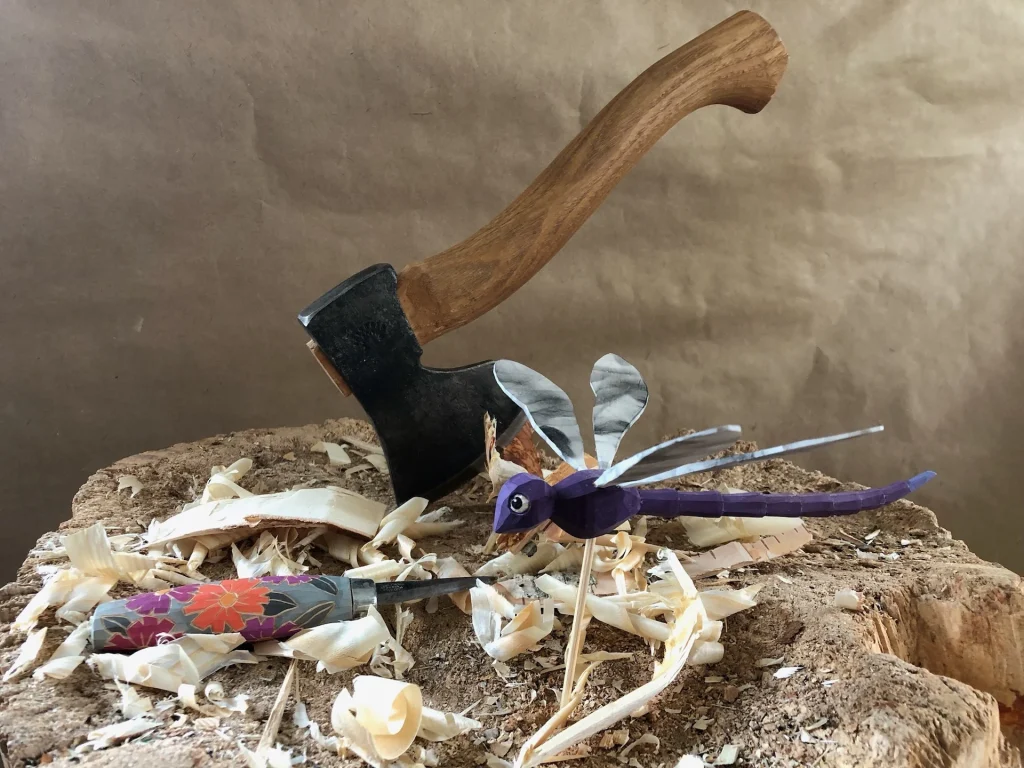 En trollslända i trä med en blommig kniv bredvid och en hammare bakom sig.